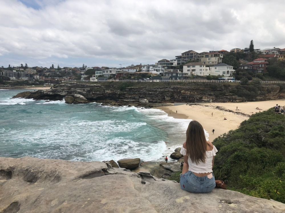 Vivir en Australia chica morena de espaldas sentada en una roca con la playa delante y el paisaje de la ciudad
